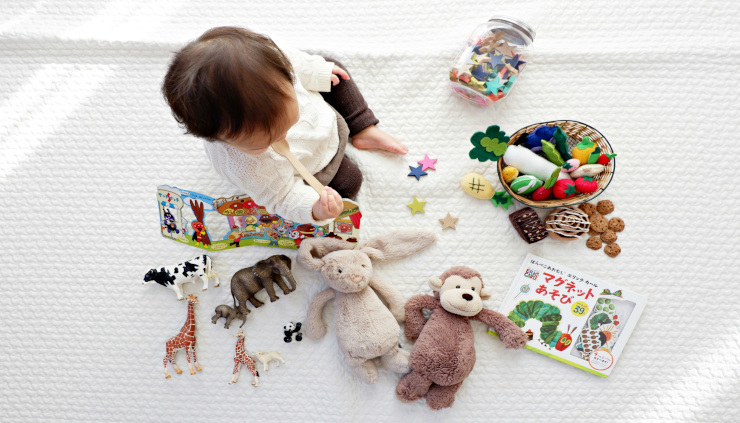 Baby sitting on a white blanket, surrounded by many toys, viewed from above.